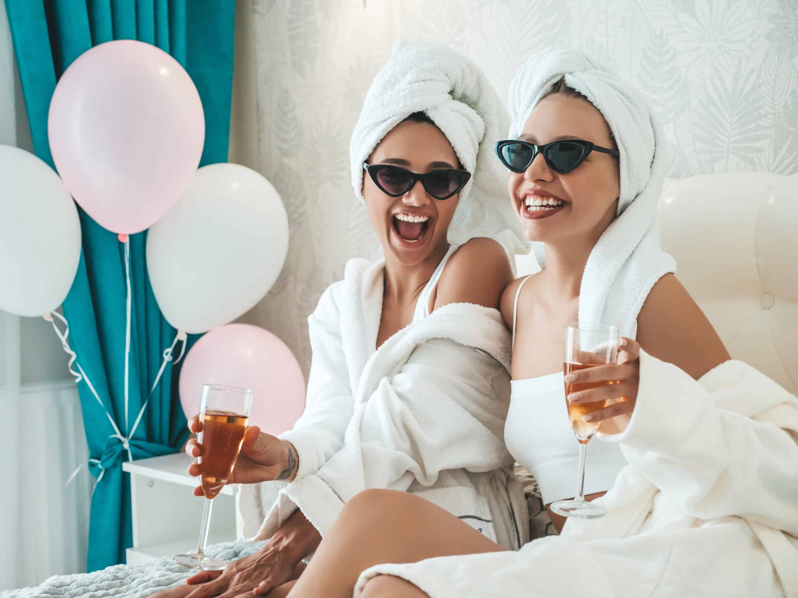 Two young beautiful smiling girls in white bathrobes and towels on head.Sexy carefree women sitting on bed in posh apartment or hotel room.Positive models drinking champagne at bachelorette party